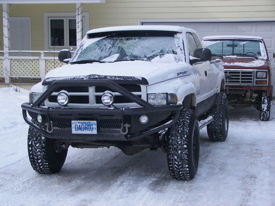 Lifted white pickup truck with custom bumper and snow on windshield, parked next to a red truck
