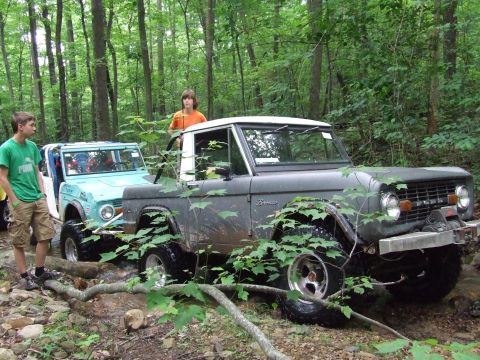 1976 Ford Bronco - Two Ford Broncos, one dark gray and one light blue, traverse a stream in a wooded area, their off-road capabilities.