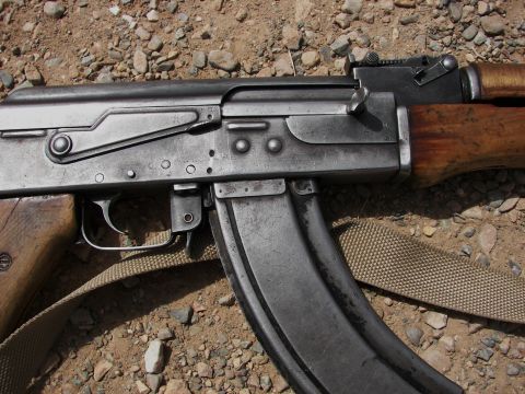 2000 Ford Crown Victoria - The receiver, magazine, and wooden furniture of an AK-47 rifle are shown in detail against a rocky ground.