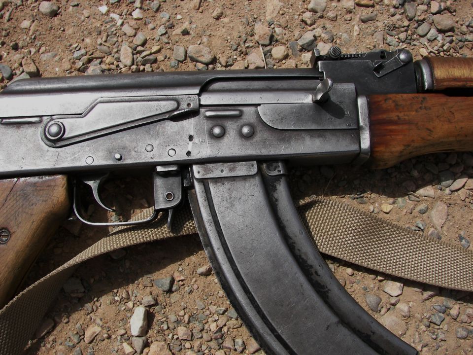 Close-up of an AK-47 rifle's receiver, magazine, and wooden stock, resting on gravel