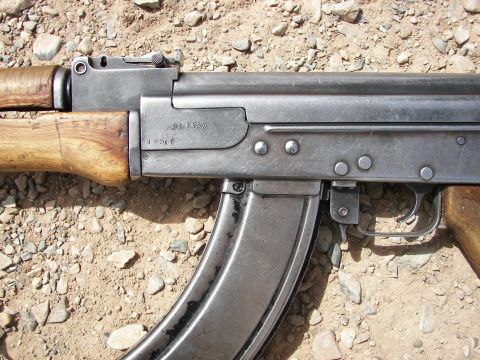 2000 Ford Crown Victoria - Detail of an AK-47 rifle receiver showing serial numbers and wooden furniture against a gravelly background.