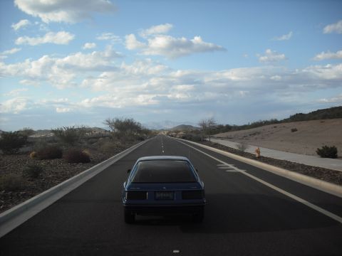 1983 Mercury Capri - A blue 1983 Mercury Capri travels down a long, straight highway through a desert environment.