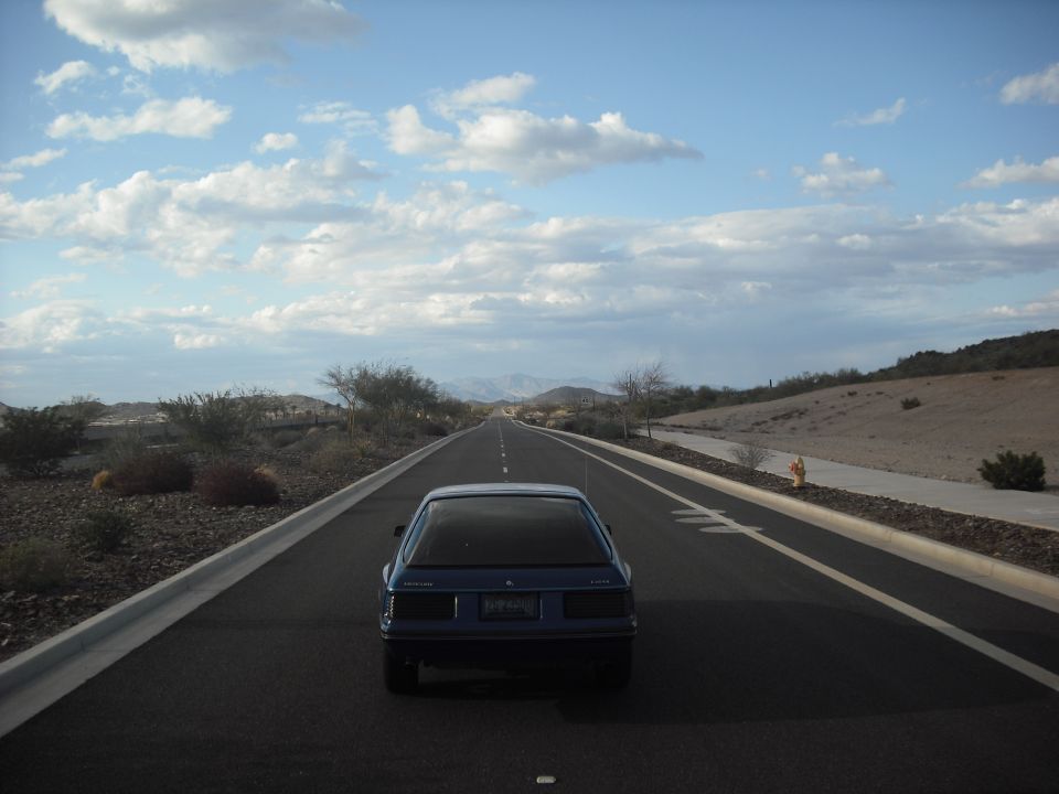 Blue 1983 Mercury Capri driving on a highway in a desert landscape