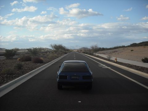 1983 Mercury Capri - A blue hatchback is seen from behind driving down a long, straight highway in a desert landscape.
