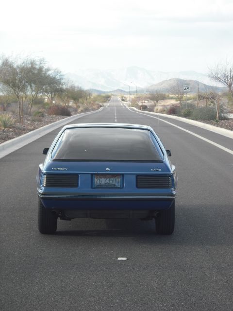 1983 Mercury Capri - A blue 1983 Mercury Capri hatchback drives down a long, straight road in a desert landscape.