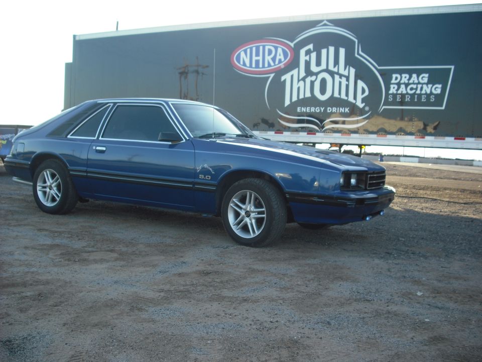 Blue 1983 Mercury Capri coupe with a 5.0 badge, parked on gravel in front of a trailer