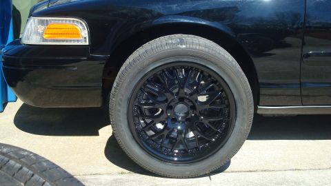 2004 Ford Crown Victoria - Close-up of a black car's front fender and wheel, showing custom rims and a portion of the headlight.