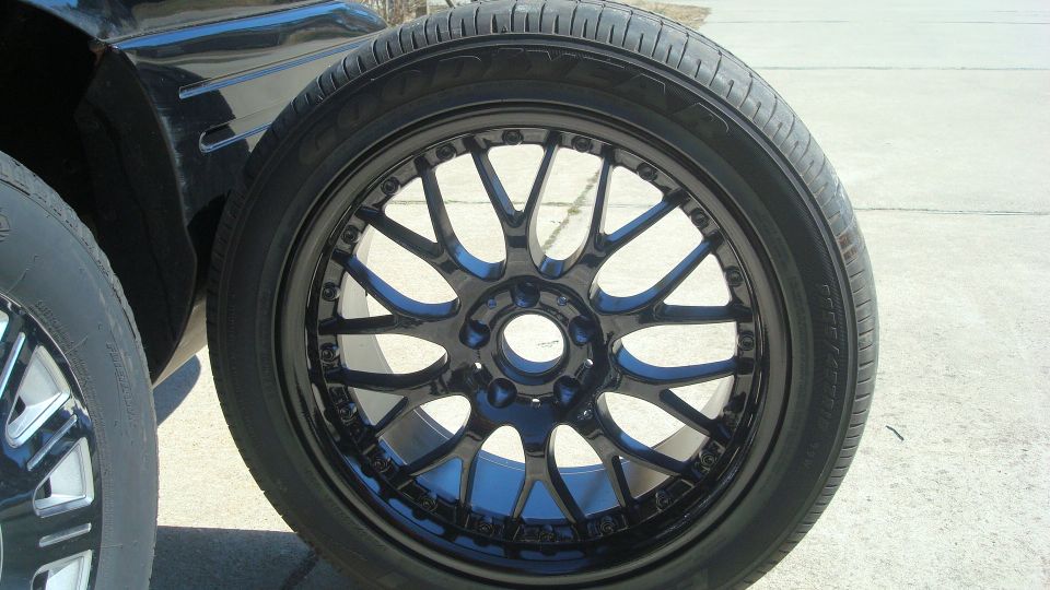 Black aftermarket wheel with a tire mounted, next to another tire. Part of a dark vehicle is visible