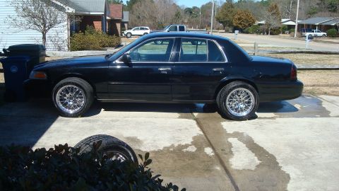 2004 Ford Crown Victoria - A black sedan is parked on a concrete driveway next to a house, showing its side profile and custom wheels.