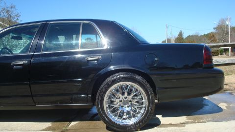2004 Ford Crown Victoria - A black sedan with custom chrome wheels is parked on wet pavement under a clear blue sky.