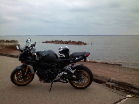 2008 Yamaha FZR1000 - A black motorcycle is parked on the side of the road with a calm body of water in the background under a cloudy sky.