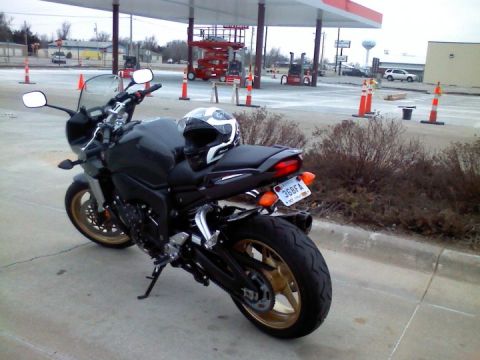 2008 Yamaha FZR1000 - A black motorcycle with gold wheels is parked at a gas station, a helmet rests on its seat.