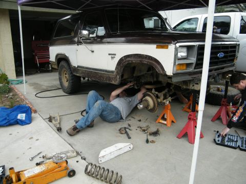 1982 Ford Bronco - Two mechanics work on the front suspension of a lifted Ford Bronco under a canopy.