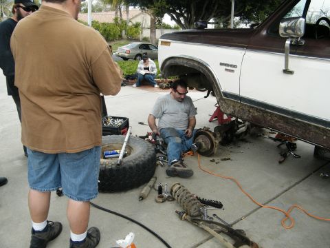 1982 Ford Bronco - A group of people are gathered around a lifted Ford Bronco undergoing maintenance, with various tools and parts laid out on the ground.