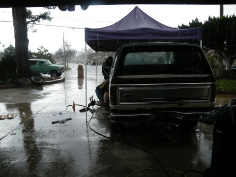 1982 Ford Bronco - A dark 1982 Ford Bronco is seen from the rear, parked under a tent during a rain shower, with a green SUV visible in the background.