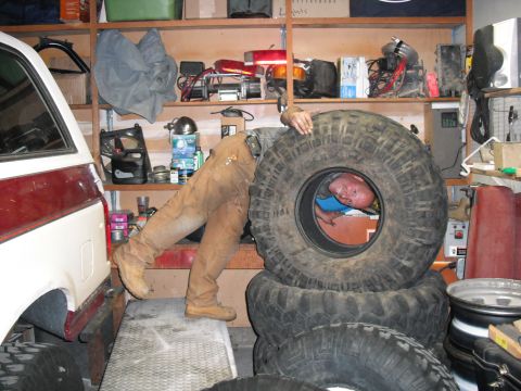 1991 Ford Bronco - A man is partially obscured by large tires next to a 1991 Ford Bronco in a cluttered garage.