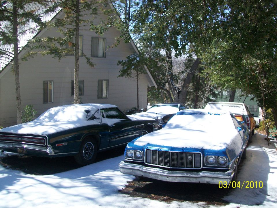 Front view of a blue 1974 Ford Ranchero GT and a dark green car covered in snow, with another vehicle behind them