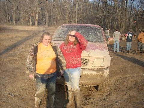 1979 Ford Bronco - Two people pose in front of a muddy red pickup truck at an outdoor event with other vehicles and people visible.