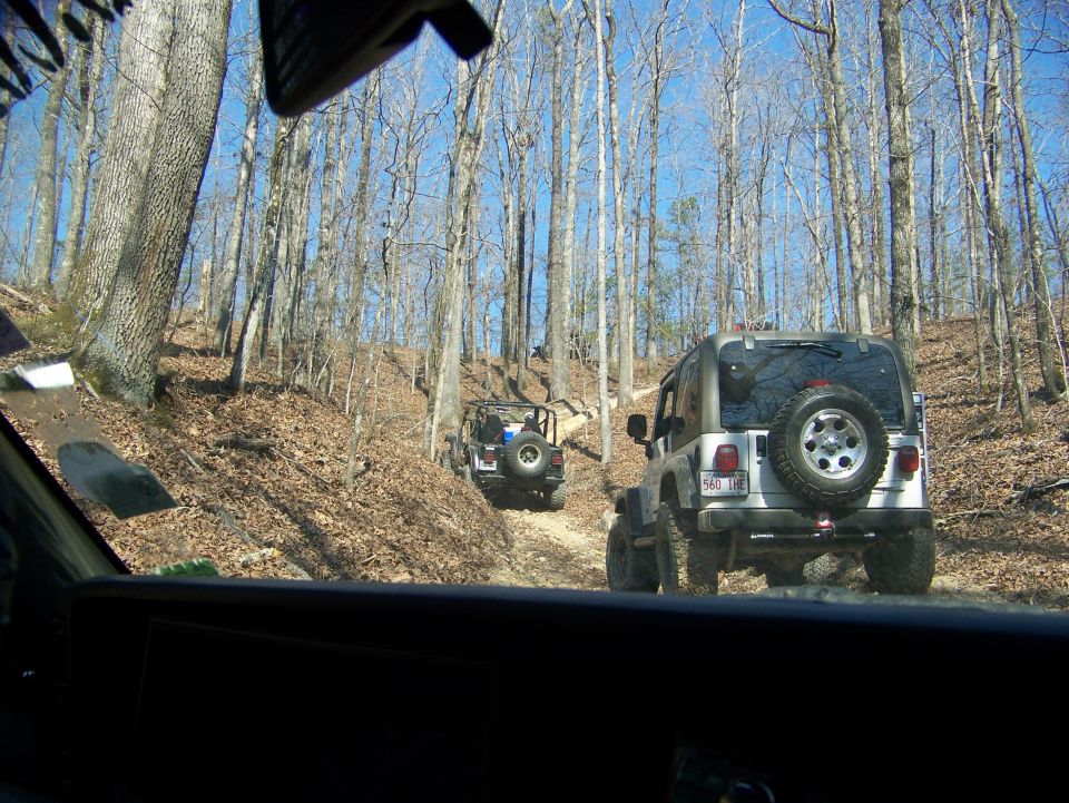 Silver Jeep Wrangler with spare tire on rear door, driving on a wooded dirt road behind another Jeep