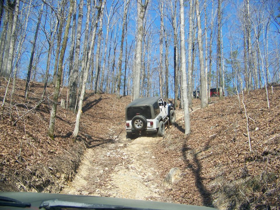 Jeep Wrangler driving up a wooded dirt trail, another vehicle visible in the distance