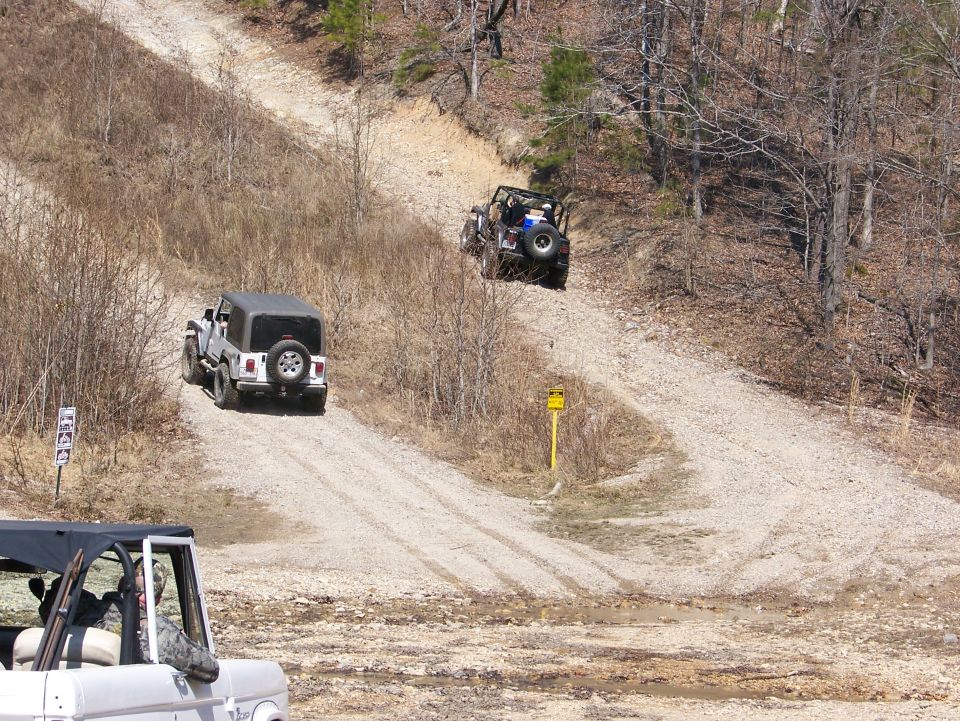 Two Jeeps driving up a steep, dry dirt trail with trees on either side. A third vehicle is partially visible in the foreground