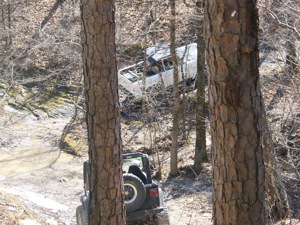 White SUV tilted on a wooded trail with a Jeep in the foreground