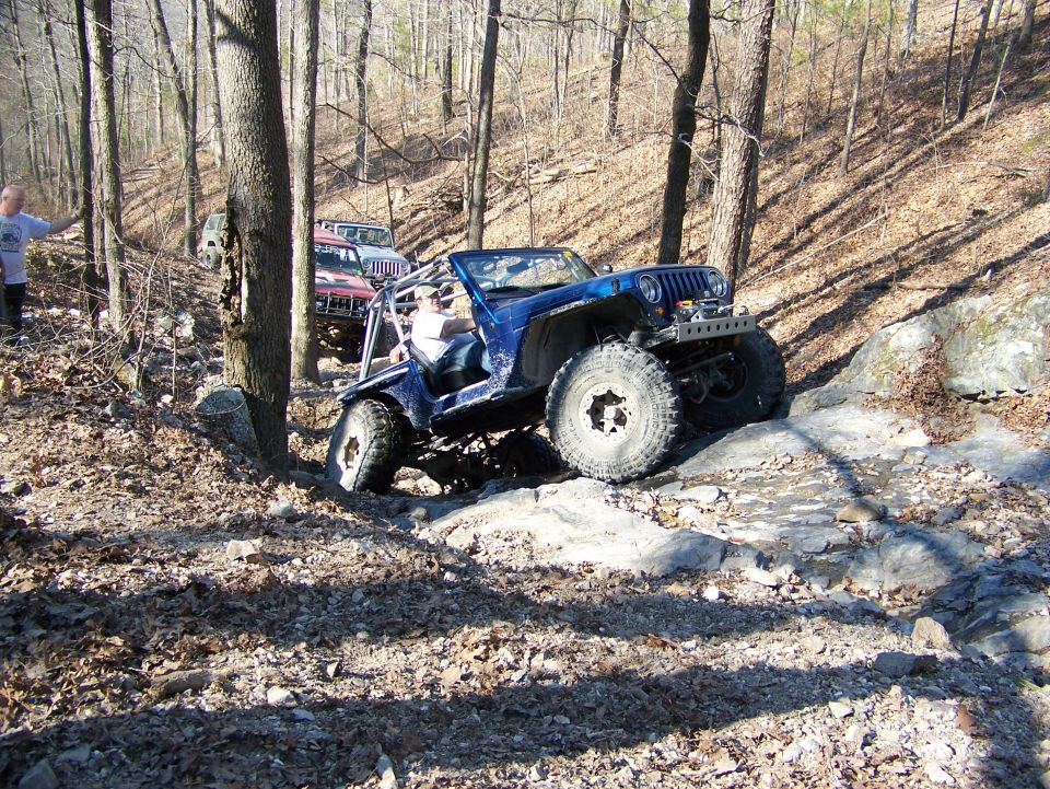 Blue Jeep navigating a rocky, wooded trail with other Jeeps behind it