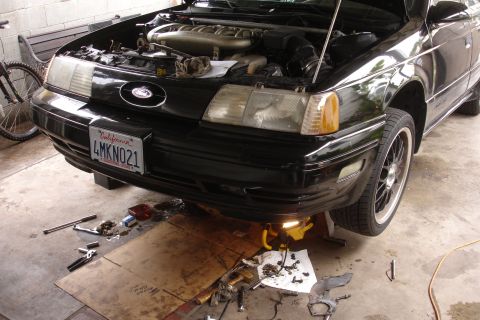 1993 Ford Taurus - The front of a black 1993 Ford Taurus is shown with its hood up, revealing the engine bay and tools scattered on the ground.