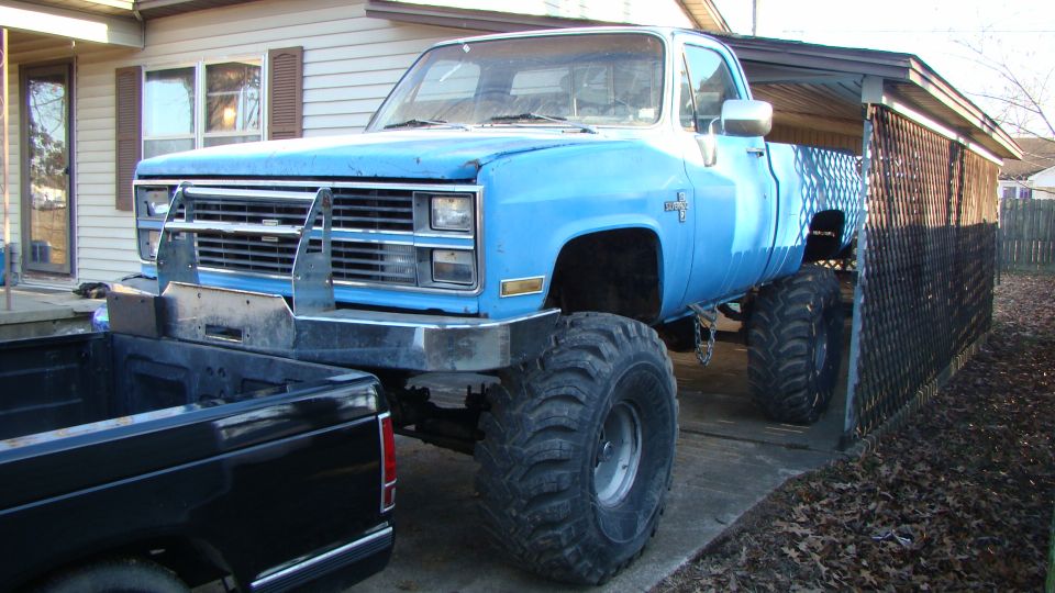 Lifted blue square-body Chevrolet pickup on mud tires beside a carport