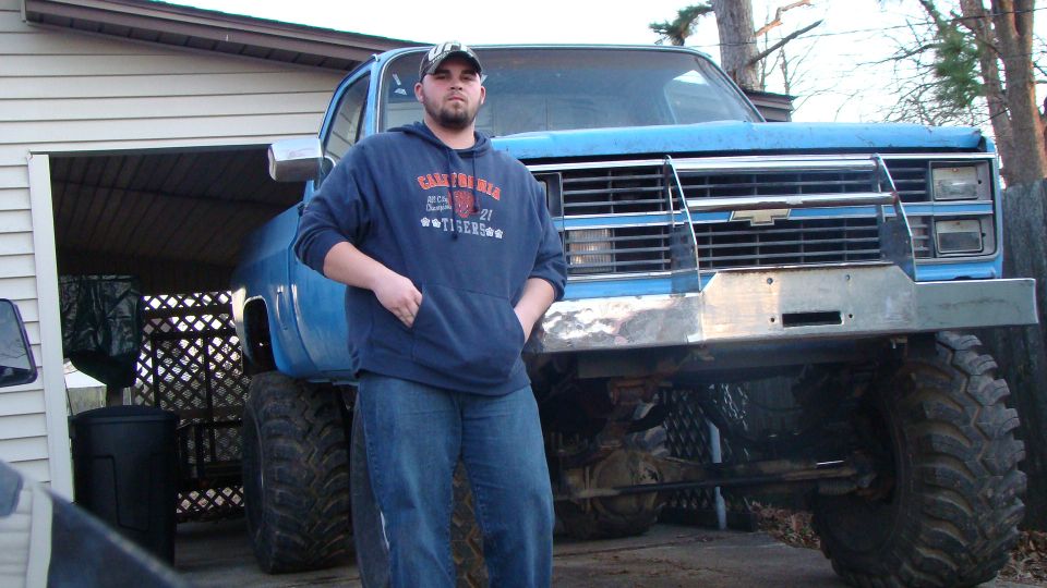 Man standing in front of lifted blue Chevy square-body pickup with big mud tires in a driveway