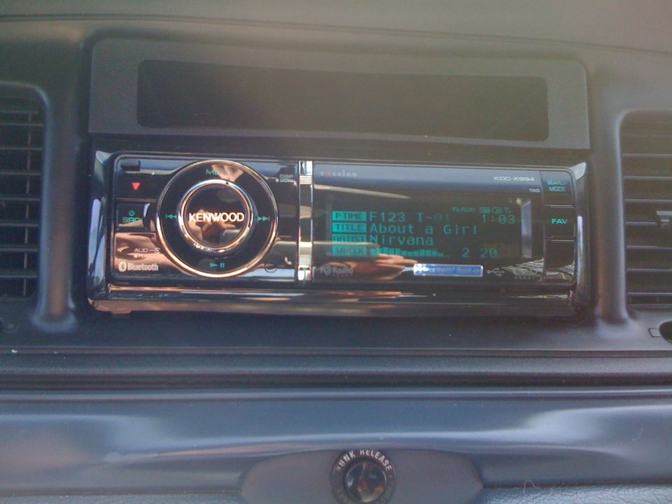 Kenwood eXcelon KDC-X994 car stereo in a gray dash displaying a Nirvana track