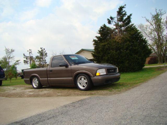 Lowered brown GMC Sonoma pickup on alloy wheels parked on gravel by a large evergreen tree