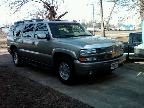 Tan Chevrolet Suburban SUV parked in a driveway, front three-quarter view with chrome grille and roof rack