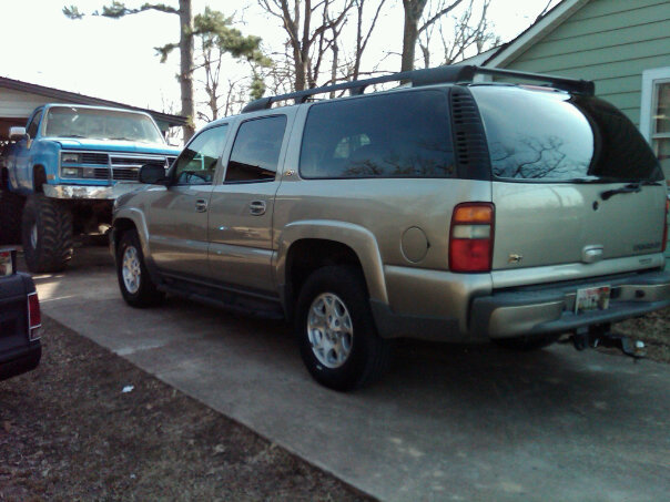 Tan Chevrolet Suburban rear three-quarter view in driveway with lifted blue Chevy pickup behind it