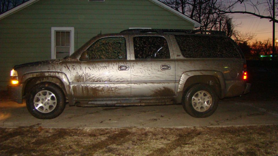 Side view of a mud-covered gray Chevrolet Suburban parked on a driveway next to a green house at dusk