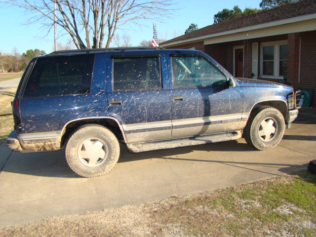 Mud-splattered dark blue GMC Yukon SUV, side view, parked in a driveway by a brick house