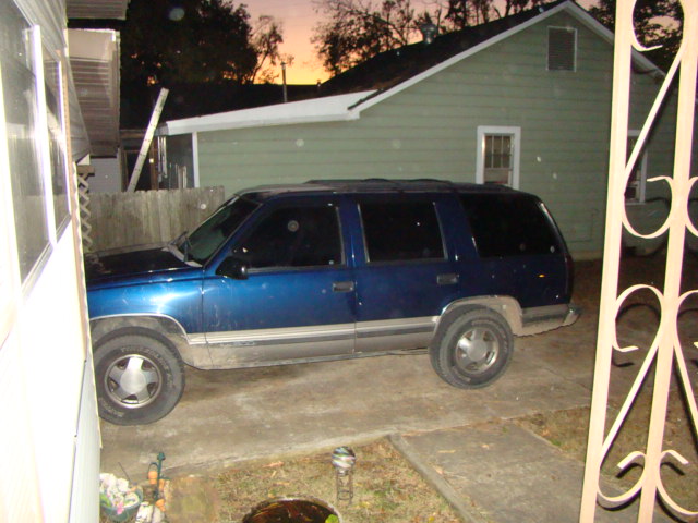 Dark blue full-size SUV parked in driveway beside a green house at dusk, side view