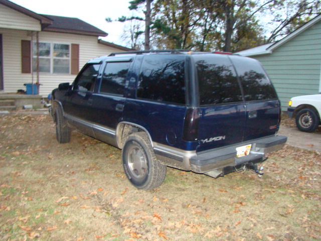 Rear three-quarter view of a dark blue GMC Yukon SUV parked on a leafy lawn in front of a house