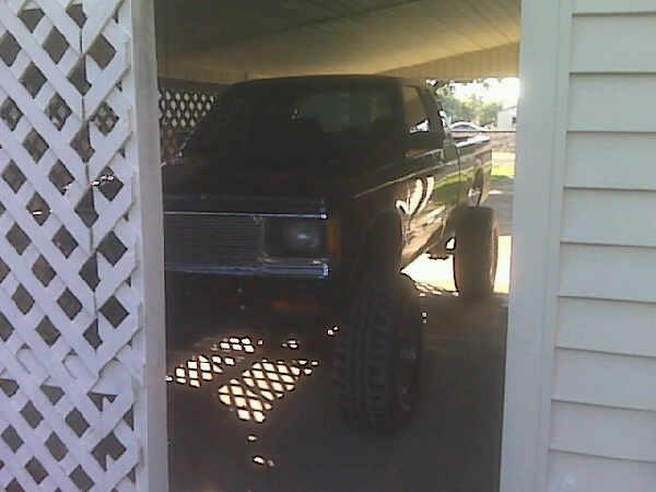 Dark lifted pickup with big off-road tires parked under a carport, seen past white lattice