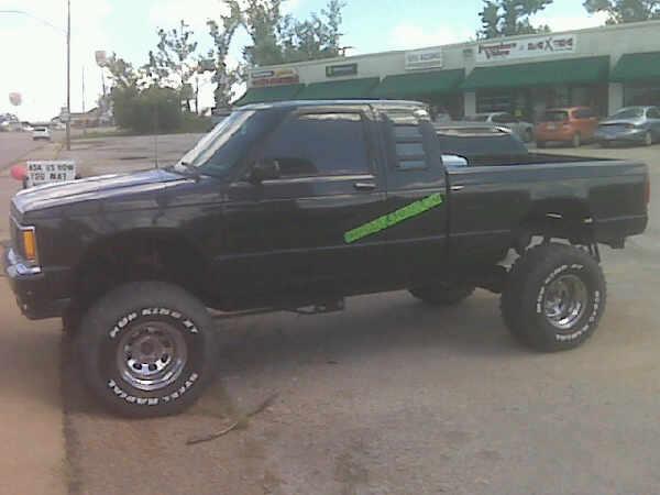 Lifted black extended-cab pickup with green door graphics, chrome wheels and white-letter mud tires, side view on street