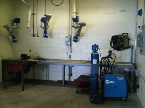 1996 Ford Bronco - A welding station is set up with a Miller welder, a gas cylinder, and a long workbench, fume extraction arms mounted on the wall.