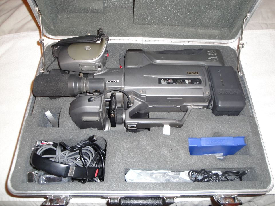 Gray Sony video camera with microphone and battery in a silver case with foam cutouts. Accessories include headphones and cables