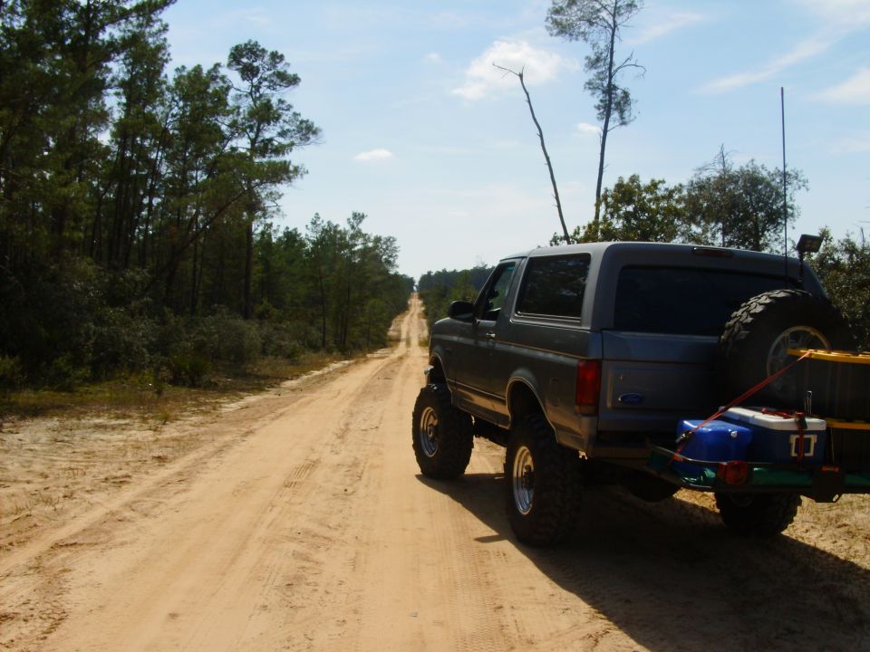 Gray 1995 Ford Bronco with spare tire on back, driving on a dirt road through trees