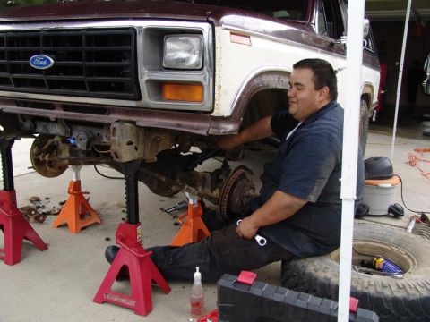 1982 Ford Bronco - A mechanic works on the front axle of a Ford truck that is supported by jack stands.