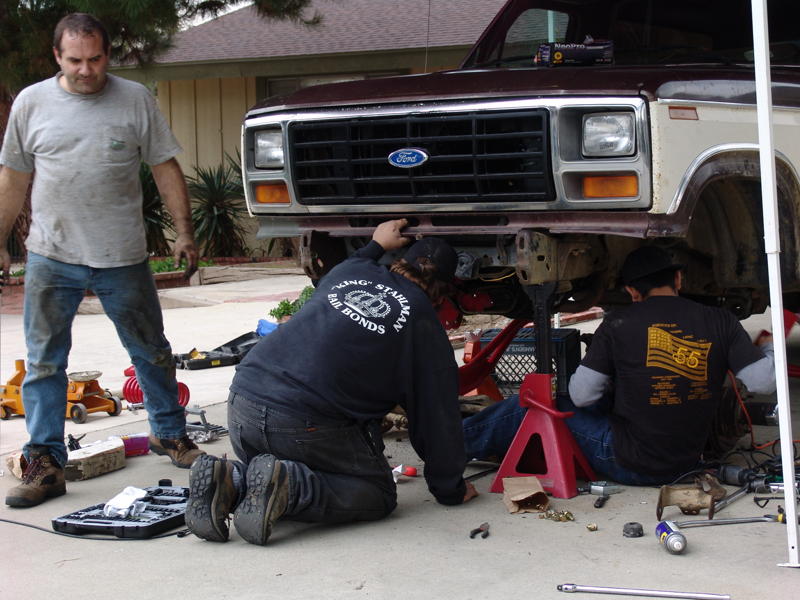 Maroon and tan Ford truck front end on jack stands, men working underneath. Visible Ford grille