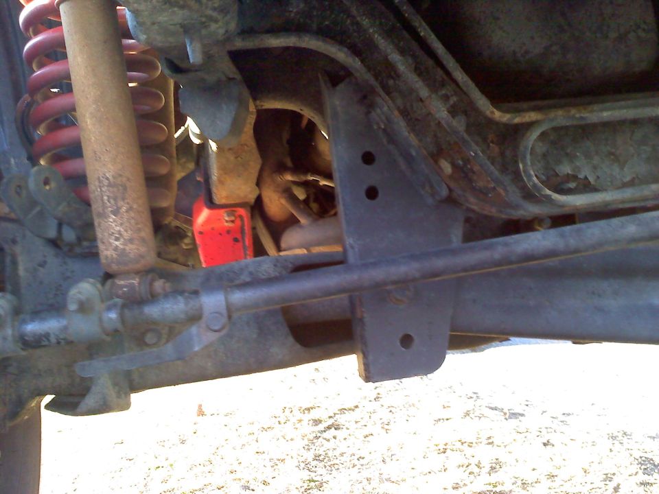 Undercarriage of a Ford Bronco showing red coil spring, shock, steering linkage, and a drilled steel bracket