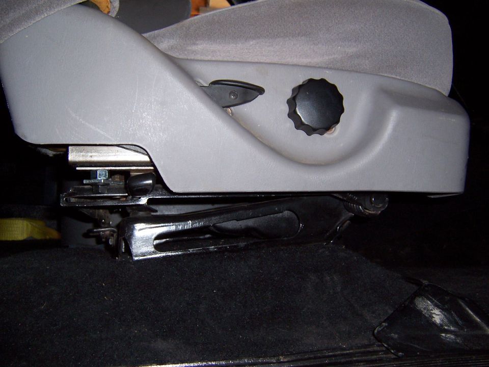 Side of gray vehicle seat showing black recliner knob, adjustment lever, and seat track mechanism over dark carpet.