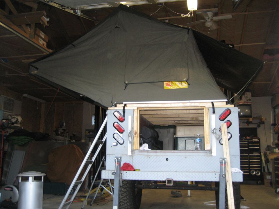 Truck bed with a large rooftop tent deployed, showing the rear of the vehicle and interior storage. Ladder attached to the side