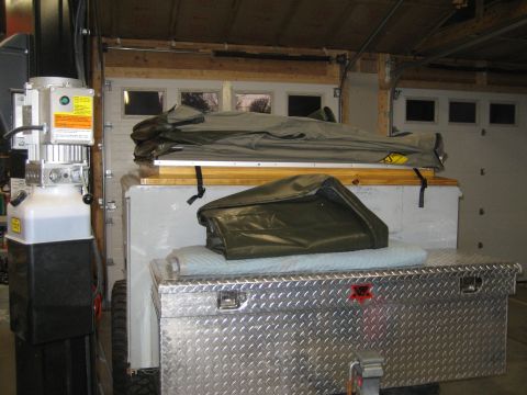1996 Ford Bronco - A trailer is parked in a garage, loaded with camping equipment including a diamond plate toolbox and folded canvas covers.