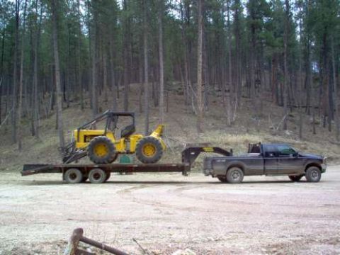 2000 Chevrolet Tahoe - A dark pickup truck tows a flatbed trailer carrying a yellow tractor through a wooded landscape.